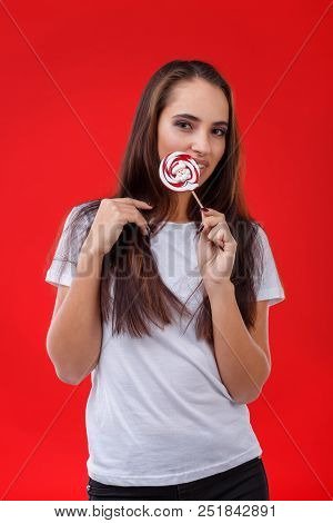 A Positive Girl With Dark Long Hair Is Licking A Sugar Candy, Posing On The Red Background.