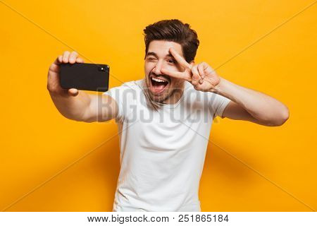 Portrait of a cheerful young man taking a sefie with mobile phone isolated over yellow background, showing peace gesture