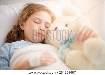 Adorable Little Girl Resting In A Hospital Bed With Her Teddy Bear. Selective Focus At Her Left Eye.