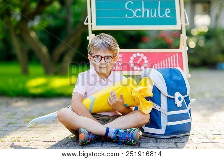 Happy Little Kid Boy With Glasses Sitting By Desk And Backpack Or Satchel. Schoolkid With Traditiona