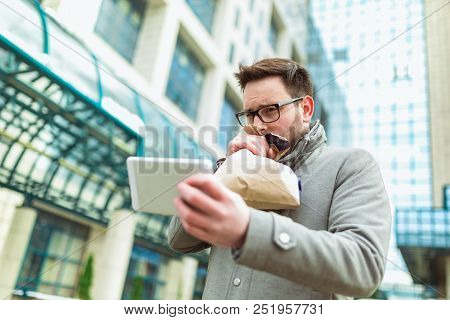 Businessman Holding Paper Bag Over Mouth As If Having A Panic Attack And Looking In Digital Tablet.