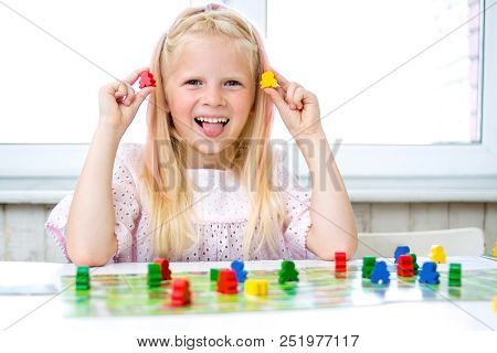 Little Blonde Girl Have Fun, Laugh And Indulge Playing Board Game. She Put Out Her Tongue. Hold Peop