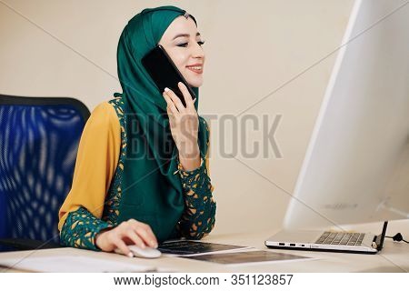 Smiling Young Beautiful Businesswoman In Traditional Hijab Working On Computer And Talking On Phone 