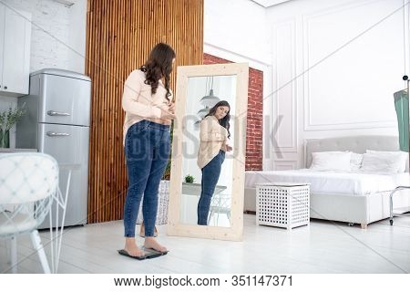 Young Woman In A Beige Blouse Standing On A Weigher And Looking At The Mirror