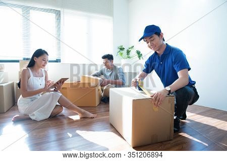 Delivery Service Worker Man In Blue Uniform Help Happy Couple Customer Packing Parcel With Tape In C