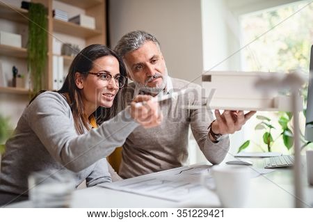 Architects With Model Of House Sitting At The Desk Indoors In Office, Working.