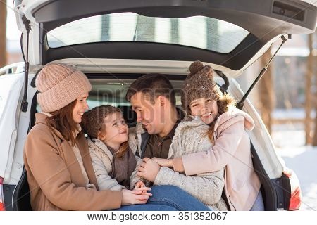 Mother, Father And Two Daughters Sitting In The Trunk Of The White Car In The Winter, Around The Tre