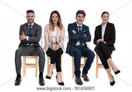 Team of 4 happy businessmen smiling and holding their arms crossed, on legs or chin while sitting on chairs on white studio background