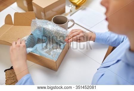 delivery, mail service, people and shipment concept - close up of woman packing mug into parcel box and it wrapping into protective bubble wrap at post office