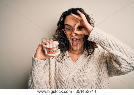 Young beautiful woman with curly hair holding plastic teeth with dental baces with happy face smiling doing ok sign with hand on eye looking through fingers