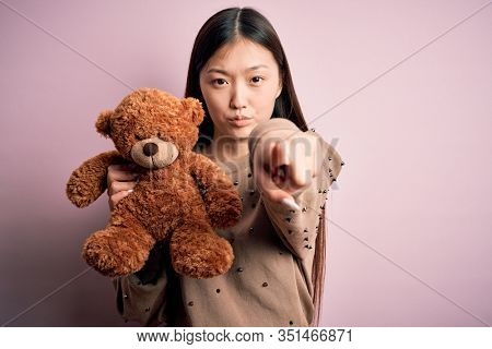 Young asian woman hugging teddy bear stuffed animal over pink isolated background pointing with finger to the camera and to you, hand sign, positive and confident gesture from the front