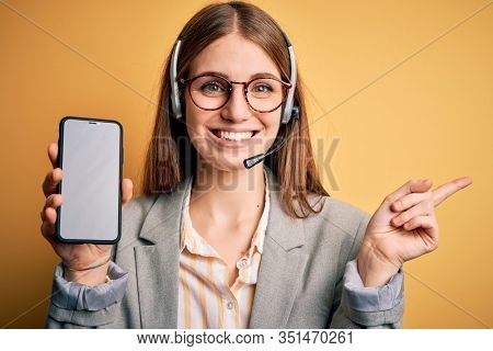 Young redhead call center agent woman using headset holding showing screen of smartphone very happy pointing with hand and finger to the side