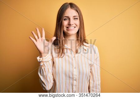 Young beautiful redhead woman wearing casual striped shirt over isolated yellow background showing and pointing up with fingers number five while smiling confident and happy.