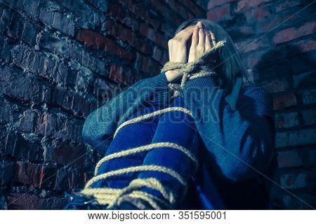 Blonde Girl With Bound Hands And Feet Is Sitting In The Basement, Covering Her Face With Her Hands A