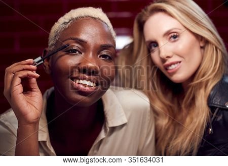 Two Young Female Friends Doing Make Up Reflected In Mirror Of Bathroom In Club