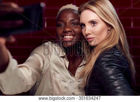 Two Young Female Friends Posing For Selfie Reflected In Mirror Of Bathroom In Club