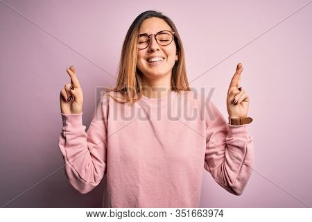 Beautiful blonde woman with blue eyes wearing sweater and glasses over pink background gesturing finger crossed smiling with hope and eyes closed. Luck and superstitious concept.