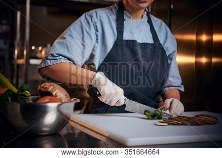 Chef Female Standing On A Dark Restaurant Kitchen, Wearing Gloves, Apron And Denim Shirt, Cutting Ve