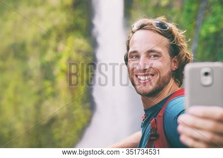 Selfie travel tourist man taking photo with phone on adventure hike vlogging for online social media influencer. Hawaii waterfall sightseeing. Smiling backpacker guy using mobile on summer vacation.