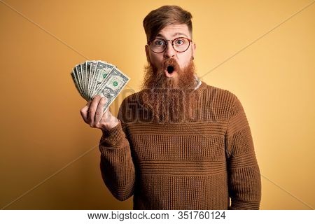 Redhead Irish man with beard holding a bunch of 1 dolla banknotes over yellow background scared in shock with a surprise face, afraid and excited with fear expression