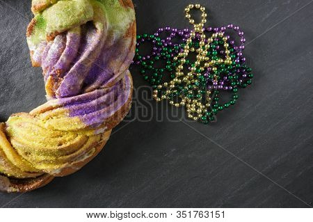 A Pile Of Green, Gold And Purple Mardi Gras Beads Next To A King Cake On A Gray Slate Background Wit