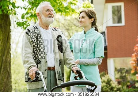 Senior Man With Walker During Walk In The Park Of Modern Nursing Home