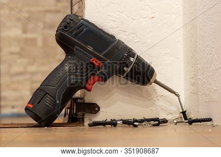 A Black Home Electric Screwdriver Leaned Against The Wall. There Are Screws Next To The Screwdriver.