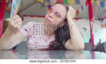 A Beautiful Young Woman Is Waving A Fan In The Sweltering Heat In A Tropical Cafe.