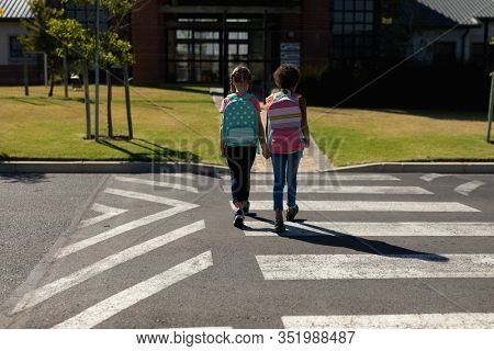 Rear view of a Caucasian and an African American schoolgirl wearing rucksacks holding hands and crossing the road at a pedestrian crossing on their way to elementary school on a sunny day