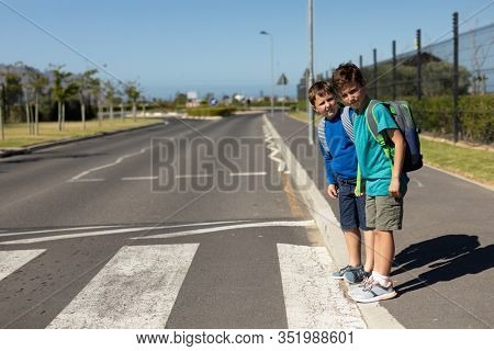 Side view of two Caucasian schoolboys looking for traffic and waiting to cross the road at a pedestrian crossing on their way to elementary school on a sunny day