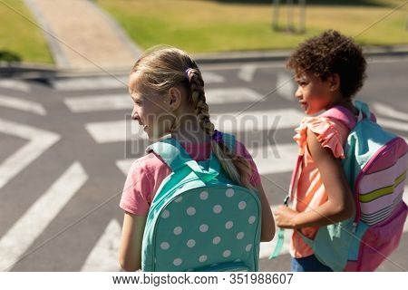 Rear view close up of a Caucasian and an African American schoolgirl wearing rucksacks looking for traffic and waiting to cross the road at a pedestrian crossing on their way to elementary school on a