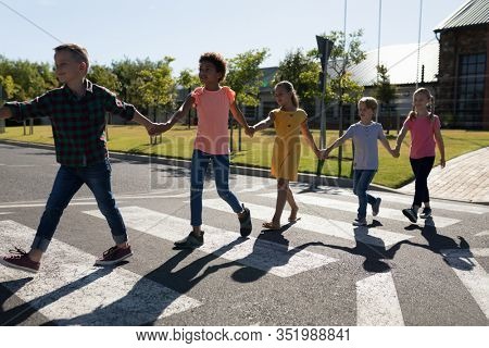 Front view of a diverse group of elementary school pupils crossing an empty road together, half way across a pedestrian crossing, holding hands on a sunny day