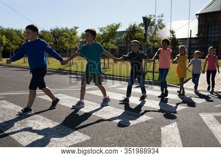 Front view of a diverse group of elementary school pupils crossing an empty road together, half way across a pedestrian crossing, holding hands on a sunny day