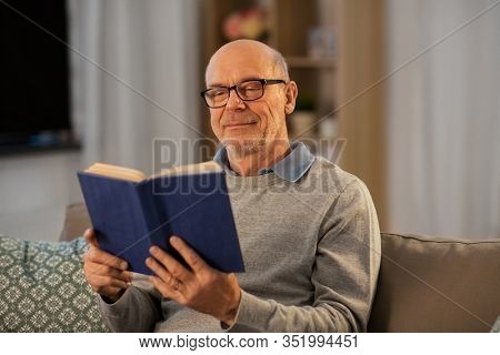 old age, leisure and people concept - happy bald senior man sitting on sofa and reading book at home in evening