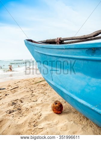 Closeup Photo Of Yellow Coconut Lying On The Ocean Baech Next To Old Wooden Fishing Boat
