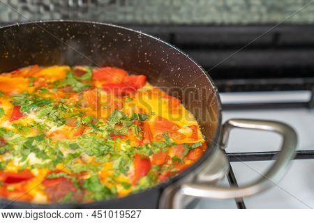 Fried Eggs With Vegetables And Herbs In Black Frying Pan On White Stove. Side View