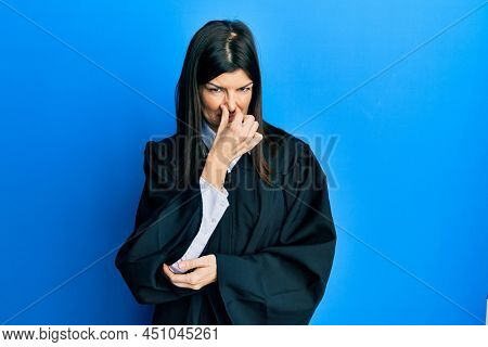 Young hispanic woman wearing judge uniform smelling something stinky and disgusting, intolerable smell, holding breath with fingers on nose. bad smell 