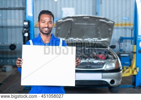 Smiling Car Mechanic In Front Broken Car At Garage Looking At Camera By Hlding Empty White Sign Boar