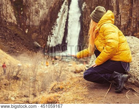 A Woman Squatting Put Her Hand Under Waterfall Flowing In The Rocks In The Distance. Optical Illusio