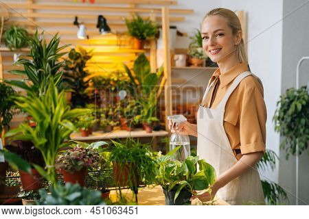 Portait Of Smiling Young Woman Florist In Apron Spraying Water On Houseplants In Pots By Sprayer, Lo
