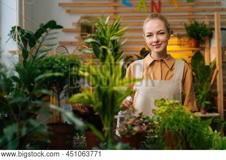 Portrait Of Attractive Young Woman In Apron Spraying Water On Houseplants In Flower Pots By Sprayer,