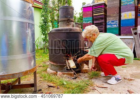 Grandma Throws Dry Branches Into The Firebox Of A Homemade Distillery Making Moonshine Schnapps, Alc