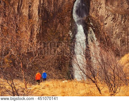 Travelers Go To A High Waterfall Flowing Between Steep Huge Rocks Among Tall Autumn Grass And Bare T