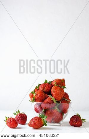 Strawberries Lie In A Glass Transparent Bowl And On A White Wooden Table On A White Background. Vert