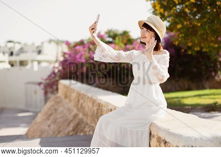A Woman In Srtaw Hat And White Dress Using Smart Phone Or Taking A Selfie On The Tropical City In Su