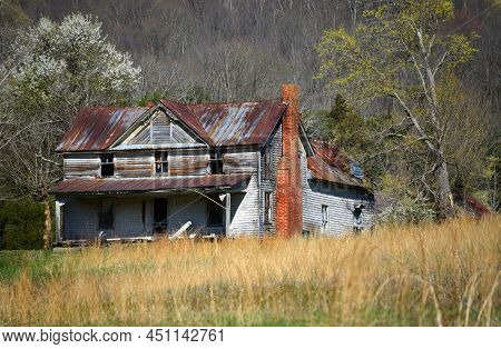 Old House Has Rusty Tin Roof And Gaping Black Glassless Windows.  Exterior Is Faded Wood And Porch I