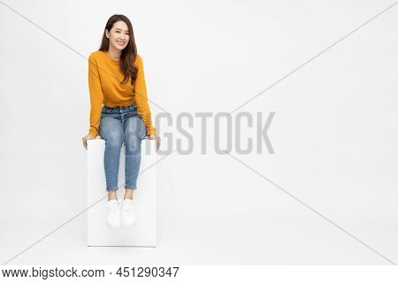 Portrait Of Young Asian Woman Sitting On White Box Isolated Over White Background, Full Body Composi