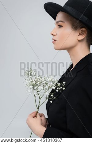 Side View Of Woman In Black Blazer And Brim Hat Near White Gypsophila Flowers Isolated On Grey