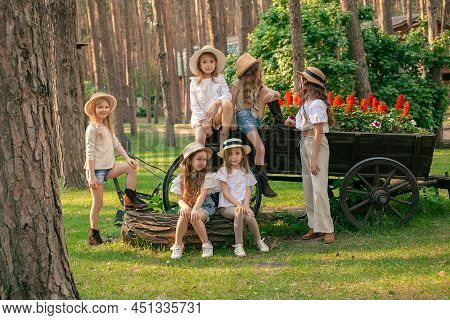 Friendly Tween Girls Posing Next To Wooden Cart-shaped Flower Bed At Country Estate