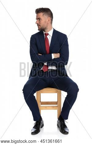 uspet businessman in suit with red tie on wooden chair crossing arms while looking to side in front of white background in studio